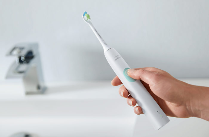 En h&aring;nd holder en hvid elektrisk Sonicare-tandb&oslash;rste fra Philips foran en h&aring;ndvask, mens tommelfingeren trykker p&aring; knappen.